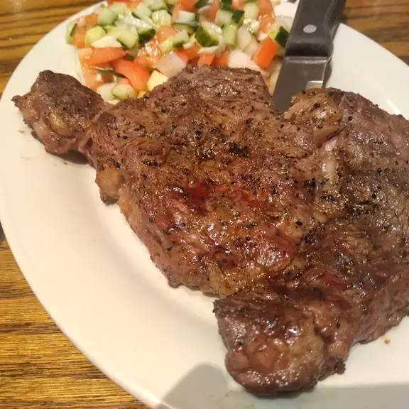 Thick flame grilled steak served on a white plate alongside a fresh Mediterranean diced salad